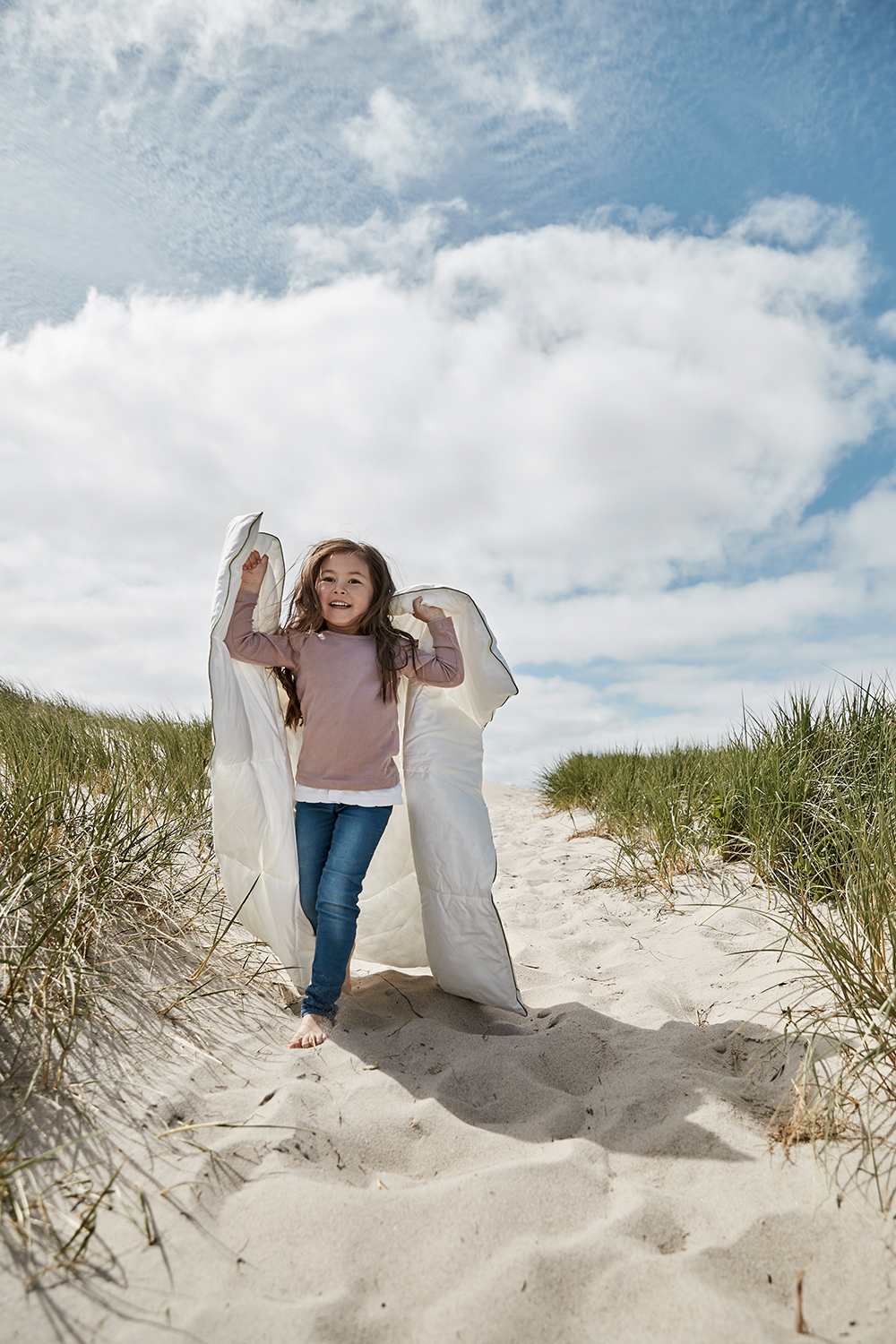 A child carrying a white duvet along a path beside the beach